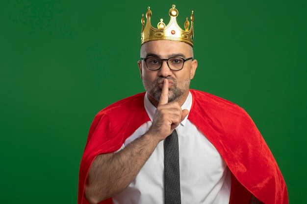 Empresario de superhéroe en capa roja y gafas con corona haciendo gesto de silencio con el dedo en los labios de pie sobre la pared verde