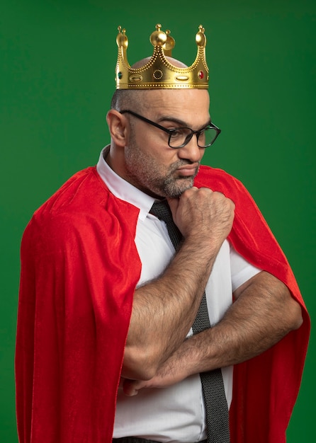 Foto gratuita empresario de superhéroe en capa roja y gafas con corona mirando hacia abajo con la mano en la barbilla con expresión seria y segura