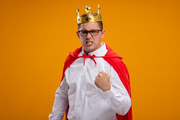 Empresario de superhéroe en capa roja y gafas con corona mirando a la cámara con expresión agresiva apretando el puño sobre fondo naranja