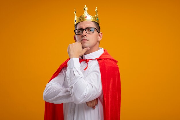 Foto gratuita empresario de superhéroe en capa roja y gafas con corona mirando a la cámara con la mano en la barbilla con expresión seria y segura de pie sobre fondo naranja