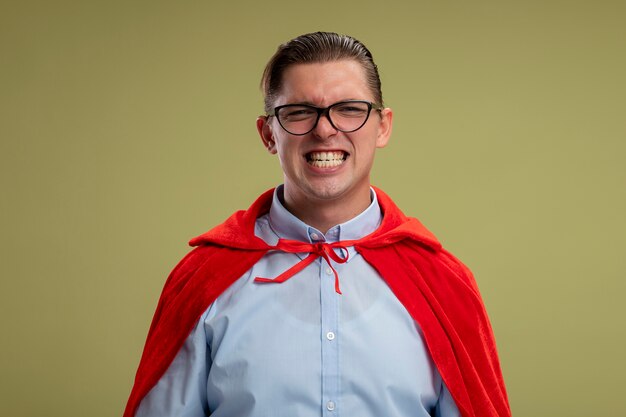 Empresario de superhéroe en capa roja y gafas mirando a la cámara sonriendo ampliamente mostrando sus dientes sobre fondo claro