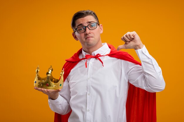 Empresario de superhéroe en capa roja y gafas sosteniendo corona apuntando a sí mismo sonriendo confiado de pie sobre la pared naranja