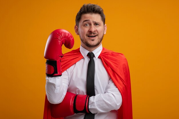 Empresario de superhéroe en capa roja y guantes de boxeo confundidos con la mano levantada de pie sobre la pared naranja