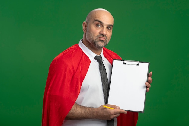 Foto gratuita empresario de superhéroe en capa roja mostrando portapapeles con páginas en blanco con cara seria