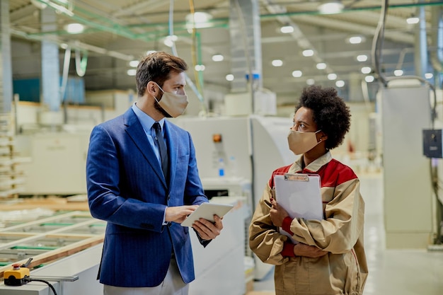 Empresario y trabajadora con máscaras faciales mientras hablan en la fábrica de carpintería