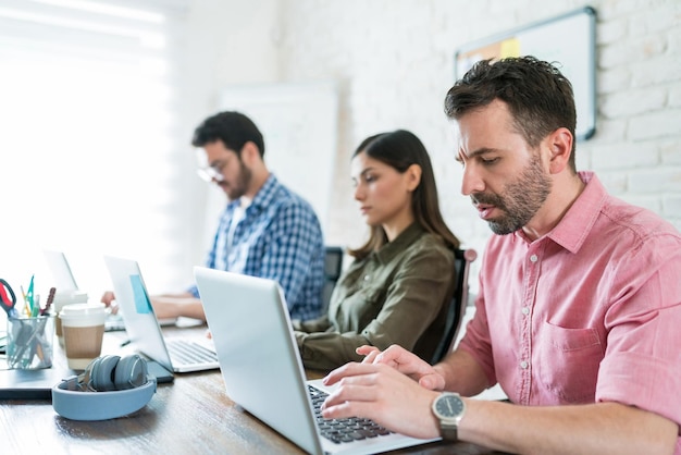 Empresario usando una computadora portátil mientras está sentado con jóvenes compañeros de trabajo en la mesa de conferencias en la oficina de coworking