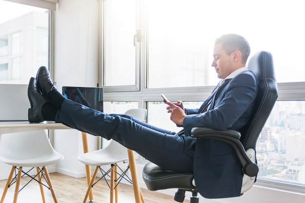 Empresario usando un teléfono celular sentado en un sillón con su pierna cruzada sobre la mesa