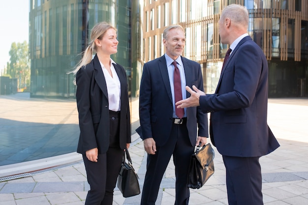 Empresarios adultos profesionales confiados que se encuentran al aire libre. Contenido hombre y mujer de negocios en traje escuchando jefe y sonriendo. Concepto de trabajo en equipo, negociación y asociación