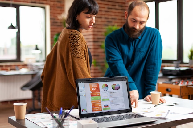 Empresarios analizando documentos de gestión discutiendo la estrategia de la empresa mientras intercambian ideas para proyectos comerciales en la oficina de inicio. Ordenador portátil con gráfico de datos de marketing en pantalla