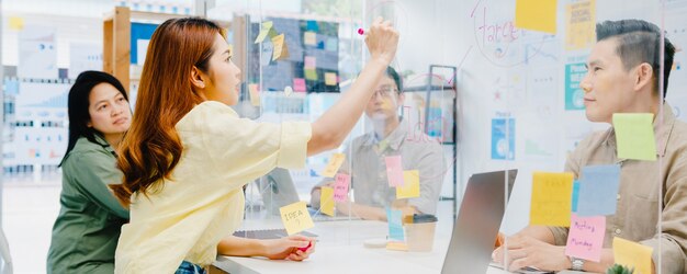 Los empresarios de Asia discutiendo una reunión de lluvia de ideas de negocios juntos comparten datos y escriben en una partición acrílica en la nueva oficina normal. Estilo de vida distanciamiento social y laboral tras coronavirus.