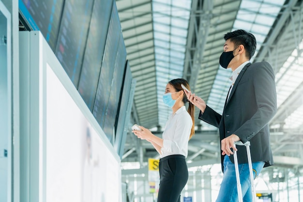 Los empresarios asiáticos usan máscara de protección facial viajes de negocios revisando el mapa y el horario de vuelo en la pantalla de embarque de información en la terminal del aeropuerto