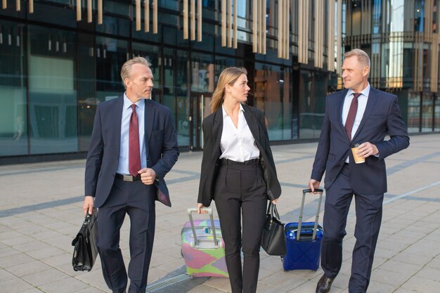 Empresarios confiados que viajan con equipaje, caminando al hotel, llevando maletas, hablando. Vista frontal. Viaje de negocios o concepto de comunicación corporativa.