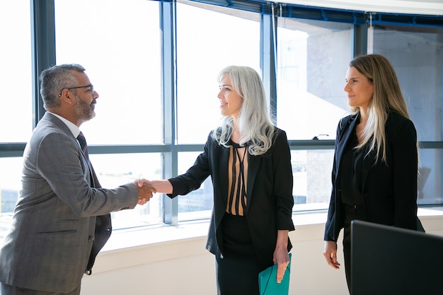 Foto gratuita empresarios de contenido saludando, reuniéndose y sonriendo. ceo indio acertado en el apretón de manos de los anteojos con la empresaria de pelo gris, hablando y mirándola. concepto de negocio y asociación