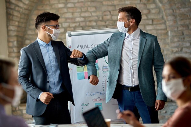 Empresarios masculinos con mascarillas protectoras golpeándose el codo mientras saludan en una reunión de negocios en la oficina