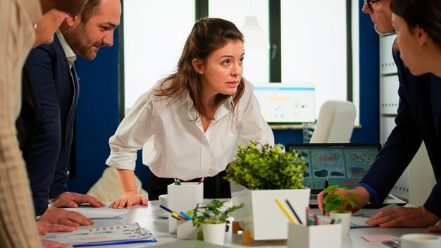 Empresarios multiétnicos analizando proyecto financiero durante la reunión corporativa. Equipo de empleados en grupo que escucha a un colega que comparte ideas discutiendo el nuevo plan de marketing comparando datos en la sala amplia.