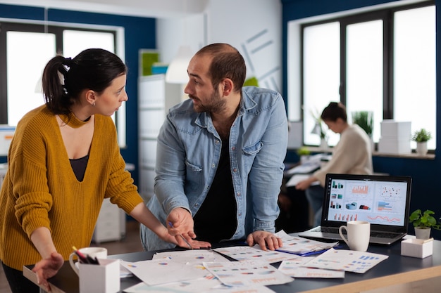 Empresarios multiétnicos intercambiando ideas sobre datos de gráficos en la oficina mirando el uno al otro. Equipo diverso de gente de negocios que analiza los informes financieros de la empresa desde la computadora. Puesta en marcha exitosa corporativa p