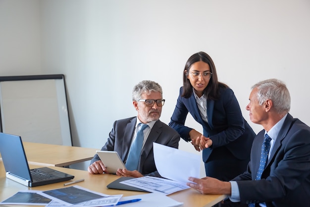 Empresarios senior y asistente joven que trabaja con estadística. Compañeros de contenido serio en trajes de oficina sentados a la mesa con portátil, documentos y tableta. Concepto de gestión y asociación