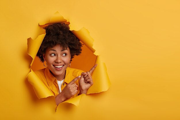 Encantada y adorable mujer con peinado afro apunta con ambos dedos índice, sonríe con alegría y muestra los dientes blancos, demuestra espacio de copia para su anuncio