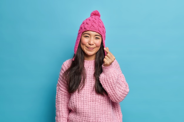 Encantada, adorable y romántica mujer asiática hace coreano como letrero, un mini corazón en la parte delantera lleva un gorro de punto y un jersey posa contra la pared azul del estudio