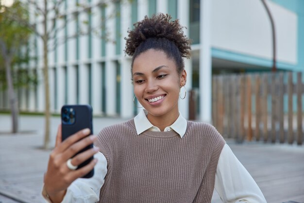 Encantada y alegre niña milenaria graba video a través de la cámara del teléfono celular comparte archivo multimedia para redes sociales toma una foto de sí misma usa conexión a Internet en itinerancia vestida con ropa elegante