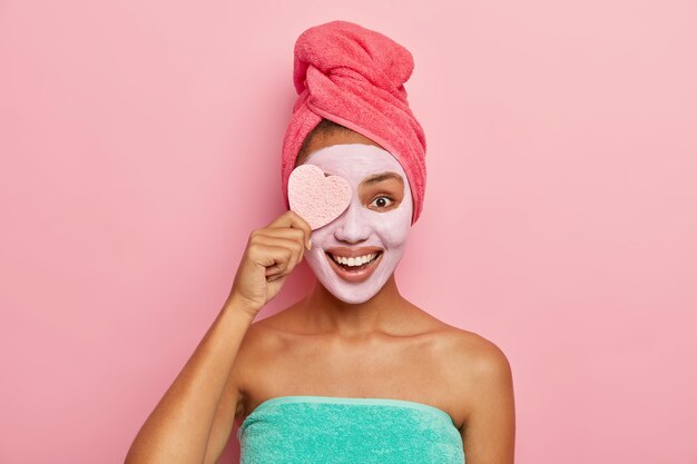 Encantada y complacida modelo femenina sostiene una esponja cosmética, aplica una mascarilla de arcilla facial que se adapta a su piel, tiene procedimientos cosméticos en el baño