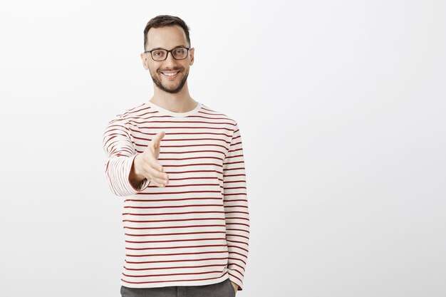 Encantada de conocerte. Retrato de hombre de negocios adulto confiado amistoso en suéter rayado, tirando de la mano hacia el apretón de manos y sonriendo con alegría, de pie sobre la pared gris