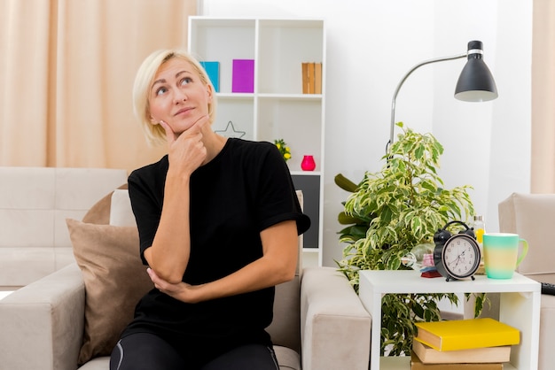 Encantada hermosa mujer rusa rubia sentada en un sillón sosteniendo la barbilla y mirando hacia arriba dentro de la sala de estar