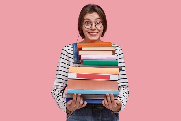 Encantada joven carga pila de libros de texto, sonríe ampliamente, aprende información útil de la enciclopedia, tiene cabello oscuro