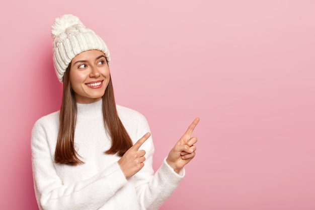 Encantada mujer de pelo oscuro se hace a un lado y señala el espacio de la copia en blanco, vestida con traje de invierno, sonríe feliz, aislado sobre fondo rosa