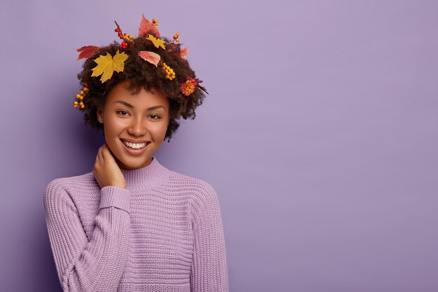 Encantada niña étnica expresa emociones sinceras, toca el cuello y se ríe alegremente, vestida con suéter de punto cálido, mira con amplia sonrisa, tiene peinado con hojas amarillas