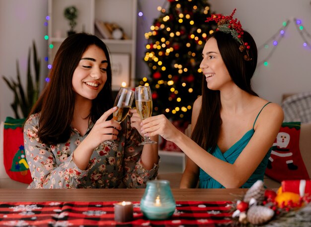 Encantado de chicas guapas tintinean copas de champán sentados a la mesa y disfrutando de la Navidad en casa
