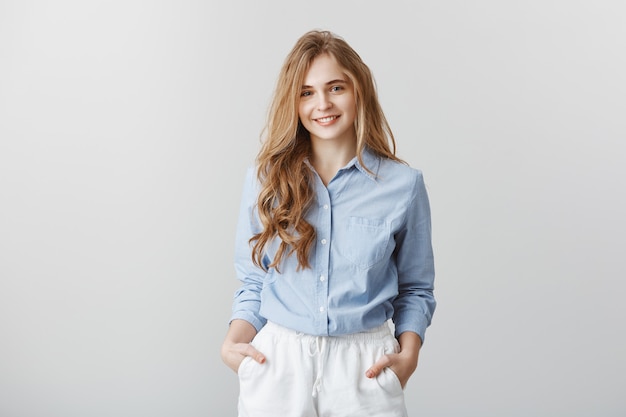 Encantado de conocer al nuevo equipo. Retrato de mujer europea atractiva de aspecto agradable con cabello rubio en blusa azul formal, tomados de la mano en los bolsillos, escuchando al cliente mientras trabaja en la oficina