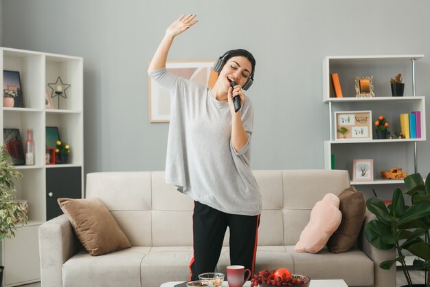 Encantado de extender la mano joven con audífonos sosteniendo el control remoto de tv canta de pie detrás de la mesa de café en la sala de estar