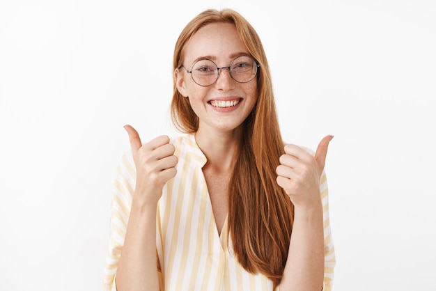 Encantado, feliz y entusiasta pelirroja creativa pelirroja con pecas lindas en gafas y blusa de rayas amarillas levantando los pulgares en señal de aprobación y gesto de acuerdo sonriendo encantada