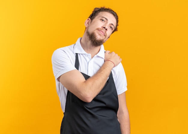 Encantado de inclinar la cabeza joven barbero vistiendo uniforme poniendo la mano sobre el hombro aislado en la pared amarilla