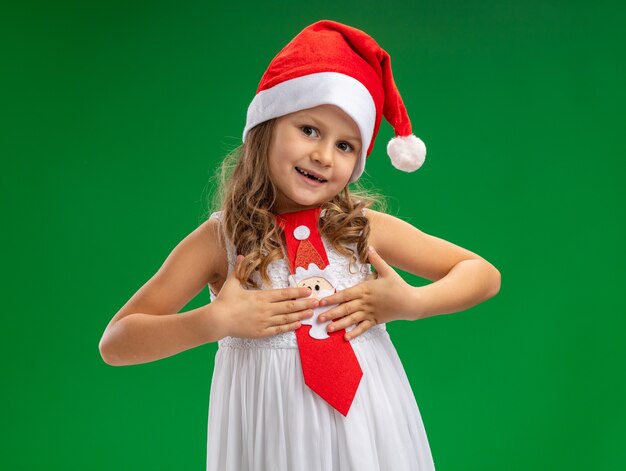 Encantado de inclinar la cabeza niña vistiendo gorro de navidad con corbata poniendo las manos sobre sí misma aislado sobre fondo verde