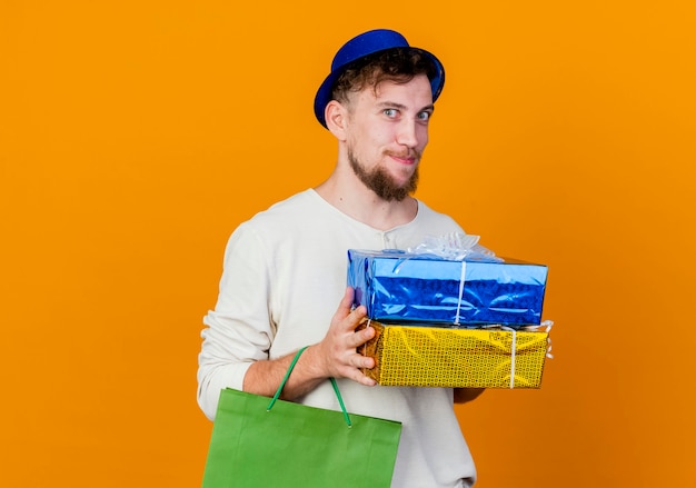 Encantado joven apuesto chico de fiesta eslavo con sombrero de fiesta sosteniendo cajas de regalo y bolsa de papel mirando a cámara aislada sobre fondo naranja con espacio de copia