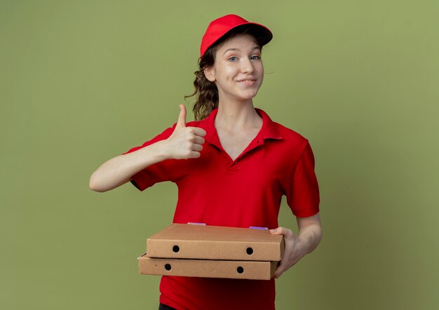 Encantado de joven bonita repartidora en uniforme rojo y gorra sosteniendo paquetes de pizza y mostrando el pulgar hacia arriba