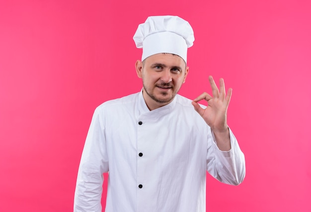 Encantado joven cocinero guapo en uniforme de chef haciendo bien signo aislado en espacio rosa