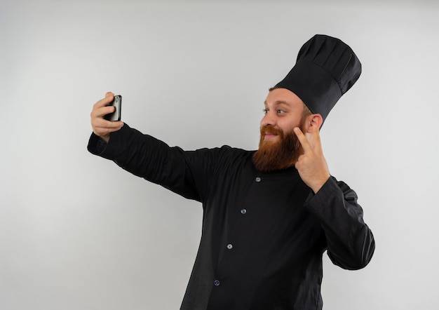 Foto gratuita encantado joven cocinero guapo en uniforme de chef haciendo el signo de la paz y tomando selfie aislado en el espacio en blanco