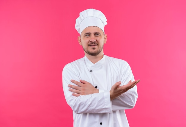 Encantado joven cocinero guapo en uniforme de chef mostrando la mano vacía y poniendo la mano en el brazo aislado en el espacio rosa