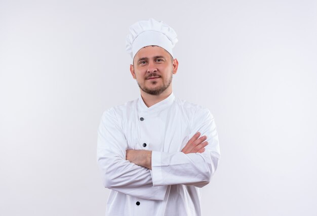 Encantado joven cocinero guapo en uniforme de chef de pie con postura cerrada aislado en el espacio en blanco