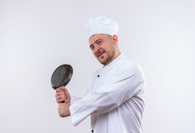 Encantado joven cocinero guapo en uniforme de chef de pie en vista de perfil sosteniendo una sartén aislado en el espacio en blanco