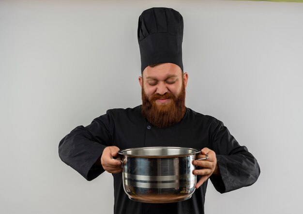 Encantado joven cocinero guapo en uniforme de chef sosteniendo y mirando la olla aislada en el espacio en blanco