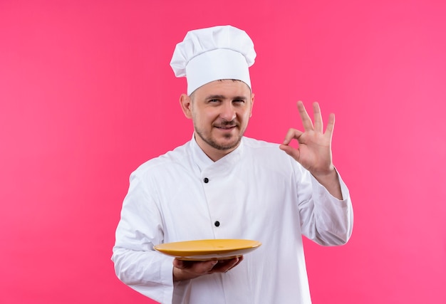 Encantado joven cocinero guapo en uniforme de chef sosteniendo la placa y haciendo bien signo aislado en el espacio rosa