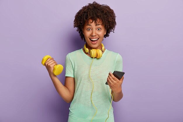 Encantado de mujer étnica charla en el teléfono móvil, escucha música durante los ejercicios de jogigng, calienta, sostiene pesas