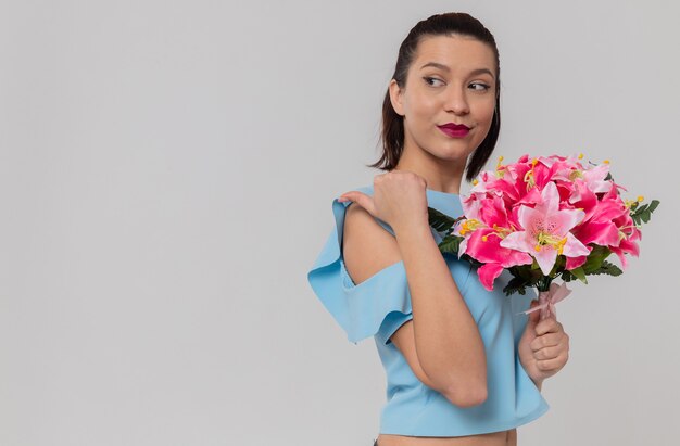 Encantado de mujer joven y bonita sosteniendo un ramo de flores y apuntando a un lado