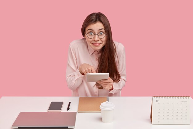 Encantado de mujer joven con gafas sostiene el panel táctil para realizar pagos en línea, vestida con una camisa elegante, posa solo en el lugar de trabajo