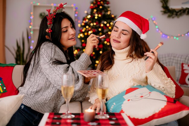 Encantado de niña bonita con corona de acebo hace maquillaje de su amiga con pincel en polvo sentado en sillones y disfrutando de la Navidad en casa