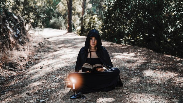 Encantador femenino con libro y candelabro sentado en el camino del bosque